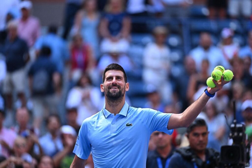 Novak Djokovic zâmbește pe terenul de la US Open, în timp ce ține în mâna stângă patru mingi de tenis, august 2025