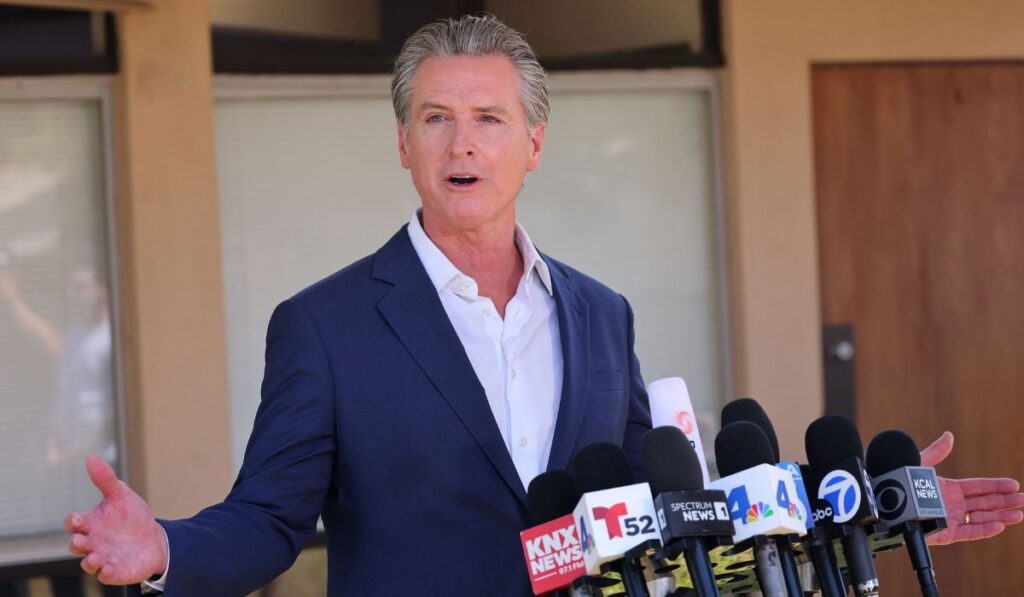 Gavin Newsom, purtând un sacou bleumarin și o cămașă bleu, ținând brațele larg deschise în cadrul unei conferințe de presă. Newsom stă în fața unui pupitru cu mai multe microfoane montate pe el