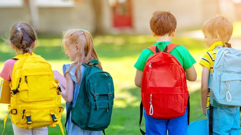 patru elevi cu ghiozdanele colorate în spate privasc spre terenul de joacă din fața lor