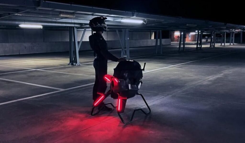 O persoană îmbrăcată în echipament de protecție pentru motociclete stă într-o cameră întunecată lângă motocicleta zburătoare Volonaut Airbike.