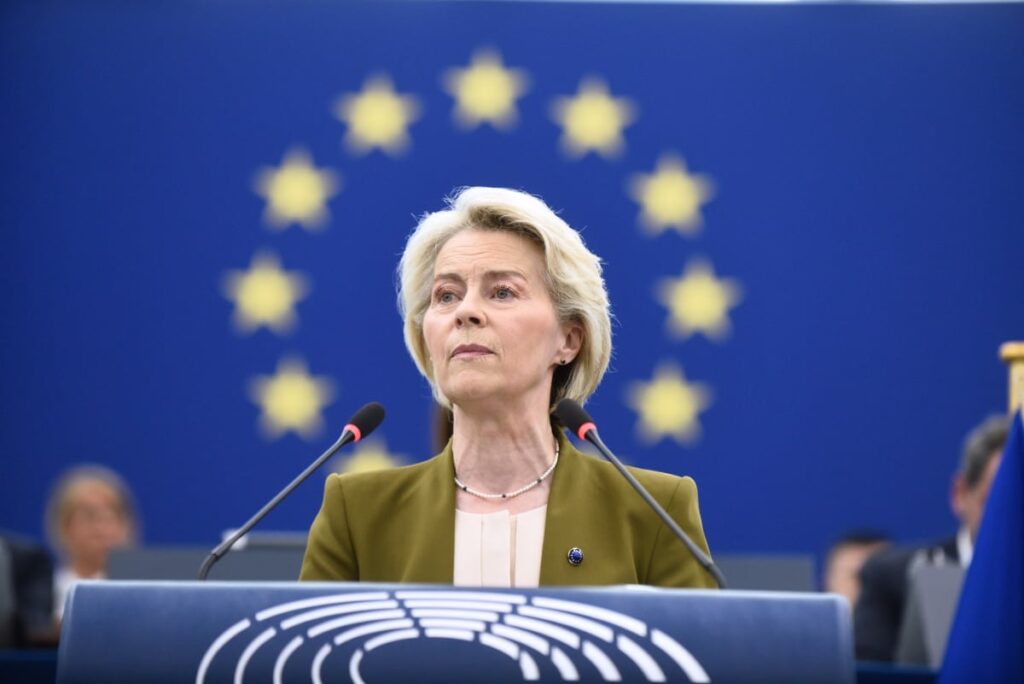 Imaginea o arată pe Ursula von der Leyen vorbind la un pupitru. Are părul scurt, blond și poartă un sacou verde-măsliniu peste un top de culoare crem. Un colier de perle este vizibil la gât. Fundalul este un steag albastru cu un cerc de stele galbene. Două microfoane se află în fața ei.