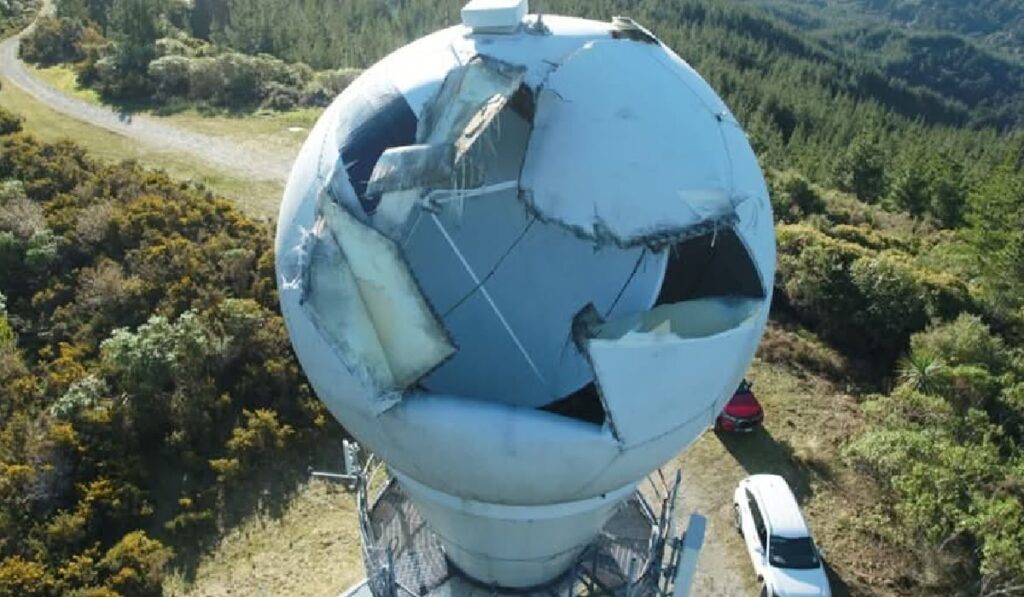 Imaginea aeriană prezintă un radom meteorologic sferic, amplasat pe un turn în vârful unui deal, care a suferit avarii majore, având panourile de protecție sparte și sfâșiate. Această structură avariată, probabil aparținând MetService New Zealand (conform numelui fișierului), este înconjurată de păduri dense și vegetație, cu două vehicule vizibile în apropiere.