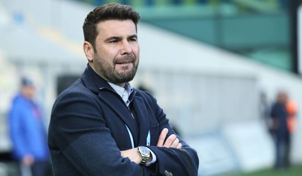 Fotbalistul Adrian Mutu, îmbrăcat la costum și cu un palton negru pe deasupra, ține mâinile încrucișate la piept și își mușcă buzele în timp ce se află pe terenul de fotbal