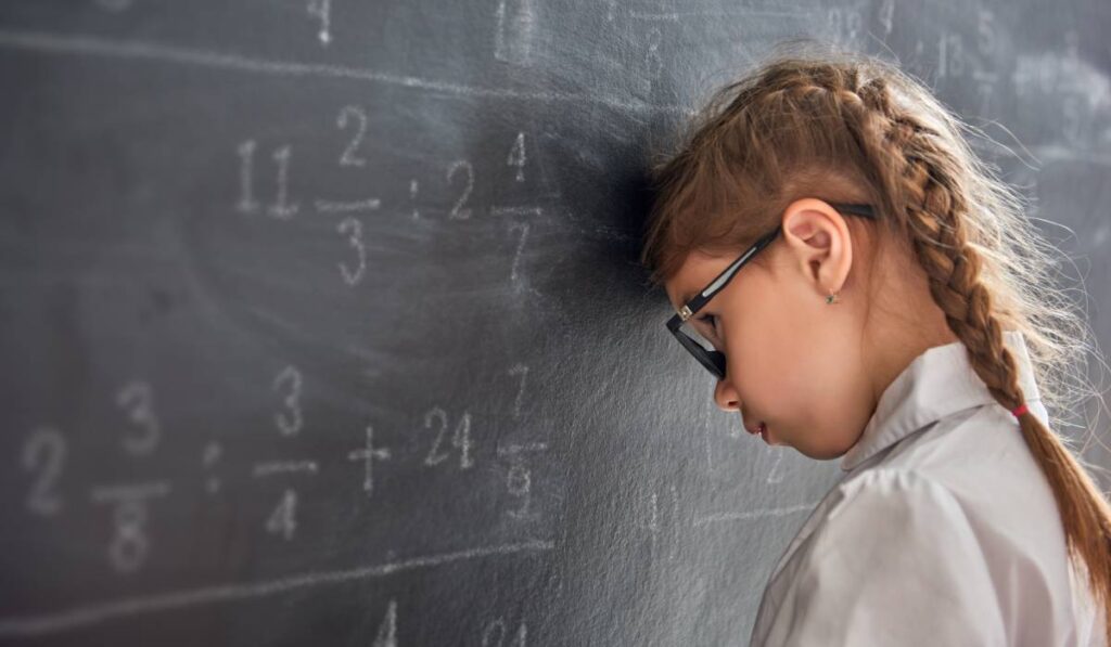O elevă blondă cu codițe împletite și ochelari, în timp ce stă rezemată cu capul de tablă și se gândește la o problemă la matematică pe care o rezolvă pentru bursa școlară