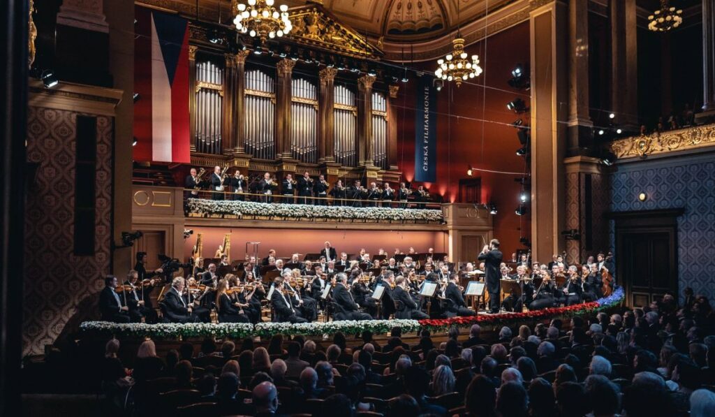 Mai mulți artiști cântă pe o scenă, în timpul Festivalului George Enescu 2025, in umnra se vede publicul, care sta pe scaune, se vad doua candelabre luminoase