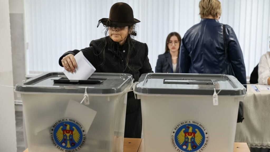 În prim-plan, o femeie în vârstă îmbrăcată în negru și cu o pălărie neagră care îi acoperă fața introduce buletinul de vot în urnă, după ce și-a exprimat opțiunea la alegerile parlamentare din Republica Moldova, iar în spate se mai văd câteva persoane, 28 septembrie 2025