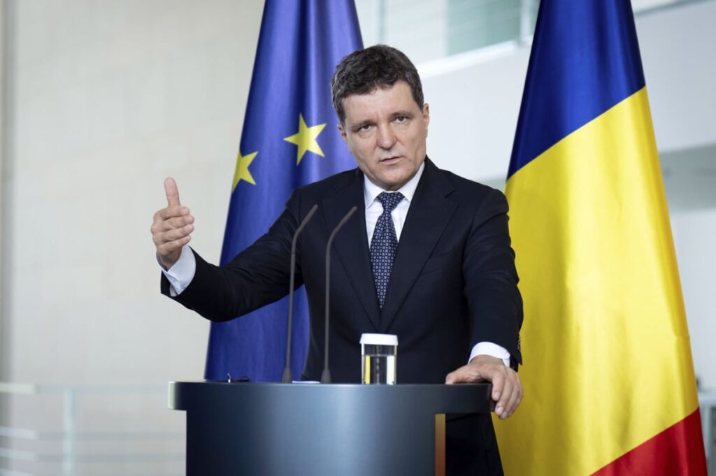 Imaginea îl arată pe Nicușor Dan, un bărbat cu părul scurt și șaten, vorbind la un pupitru. El este îmbrăcat într-un costum formal, compus dintr-un sacou închis la culoare, o cămașă albă și o cravată cu un model subtil. Într-o mână ține un microfon, iar cu cealaltă gesticulează. În spatele său, sunt două steaguri: cel al Uniunii Europene, de culoare albastră cu stele galbene, și steagul României, cu benzi verticale de culoare albastră, galbenă și roșie. Pe pupitru se află un pahar cu apă.