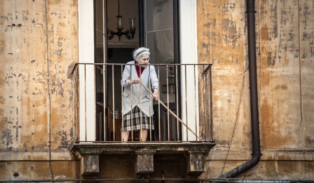 O femeie în vârstă din Italia, îmbrăcată cu o jachetă gri din lână și o fustă în carouri, în timp ce dă cu mătura pe balconul casei sale. Pereții maro ai clădirii au tencuiala căzută