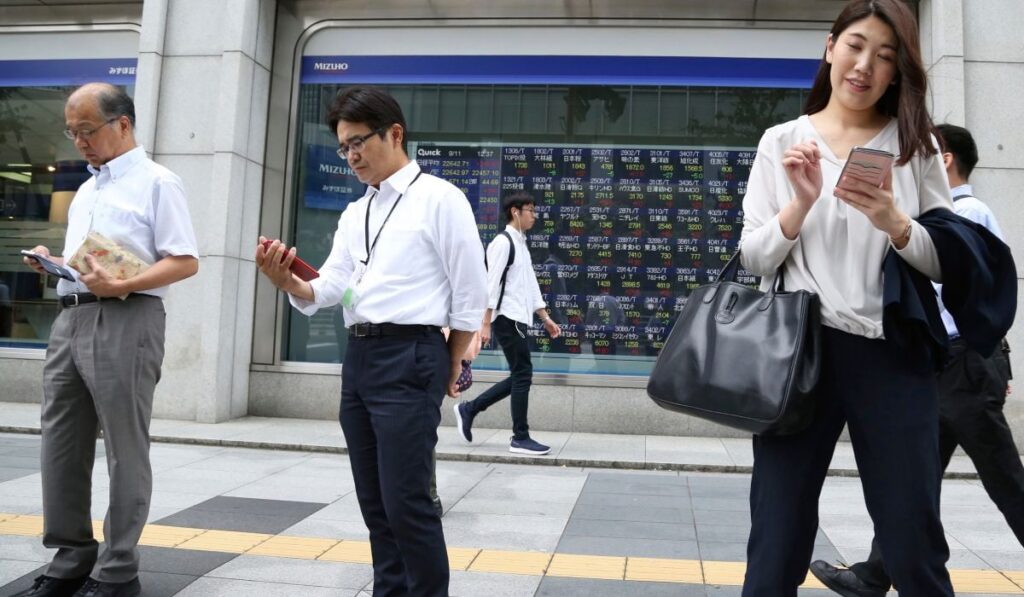 Oameni se plimbă pe stradă în Japonia, îmbrăcați business, în timp ce folosesc smartphone-uri.