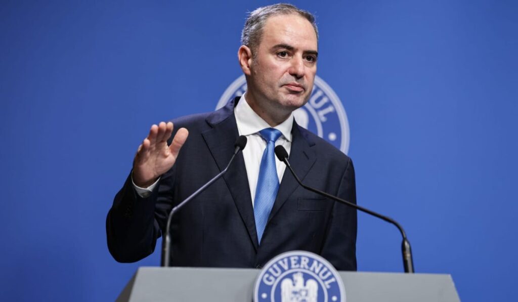 Alexandru Nazare, îmbrăcat într-un sacou negru, o cămașă albă și o cravată albastră, în timpul unei conferințe de presă de la Palatul Victoria, 13 august 2025