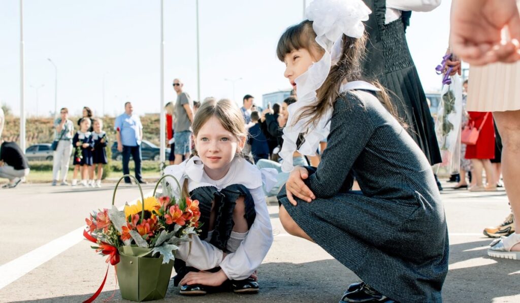 O elevă de clasa întâi, îmbrăcată în uniformă școlară. Stă pe trotuar și ține în mână un buchet de flori, langa ea o alta eleva mai mare, in uniforma cu codite cu pampoane, mai multi copii si parinti se vad in spate. Atmosferă de început de an școlar.