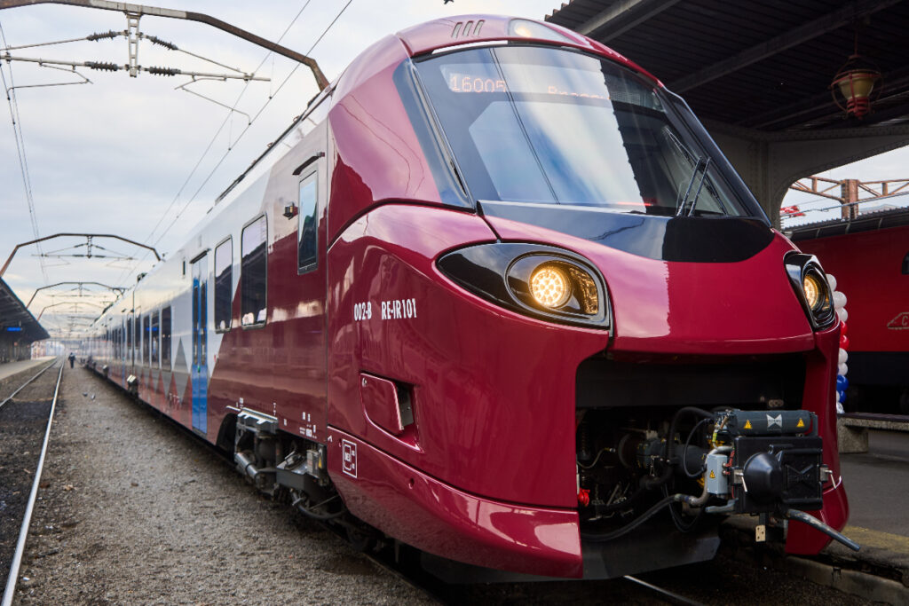 Fotografia surprinde un tren modern, de culoare roșie-grena, cu accente albe și gri, aflat într-o gară. Partea din față a locomotivei este rotunjită și are două faruri rotunde, cu lumina aprinsă. Pe laterale se pot observa vagoanele. Imaginea este realizată din unghiul șinelor, dând o senzație de viteză și dinamism.