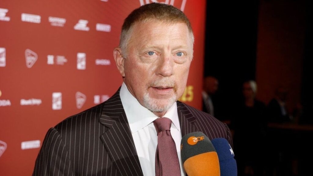 Boris Becker, 57 de ani, in costum inchis la ciloare , camasa alba si cravata maro, la decernarea premiilor Sport-Bild, Hamburg, in timpul unui interviu, cu microfonul in fata si un panou rosu cu scris alb in spate, 25 august 2025