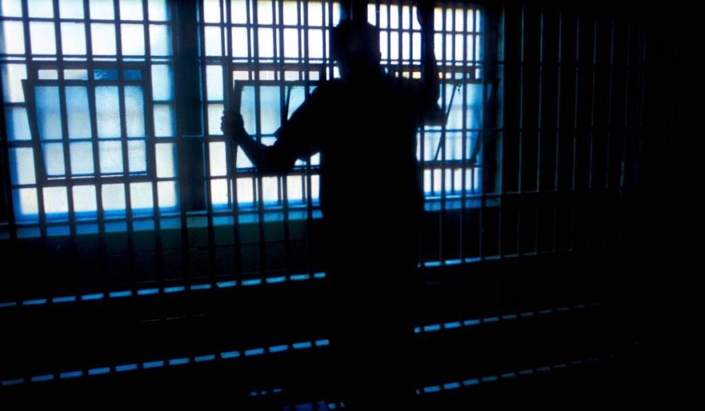 Un bărbat în închisoare, fotografiat de la spate, în timp ce privește pe geamurile cu grilajele ale pușcăriei