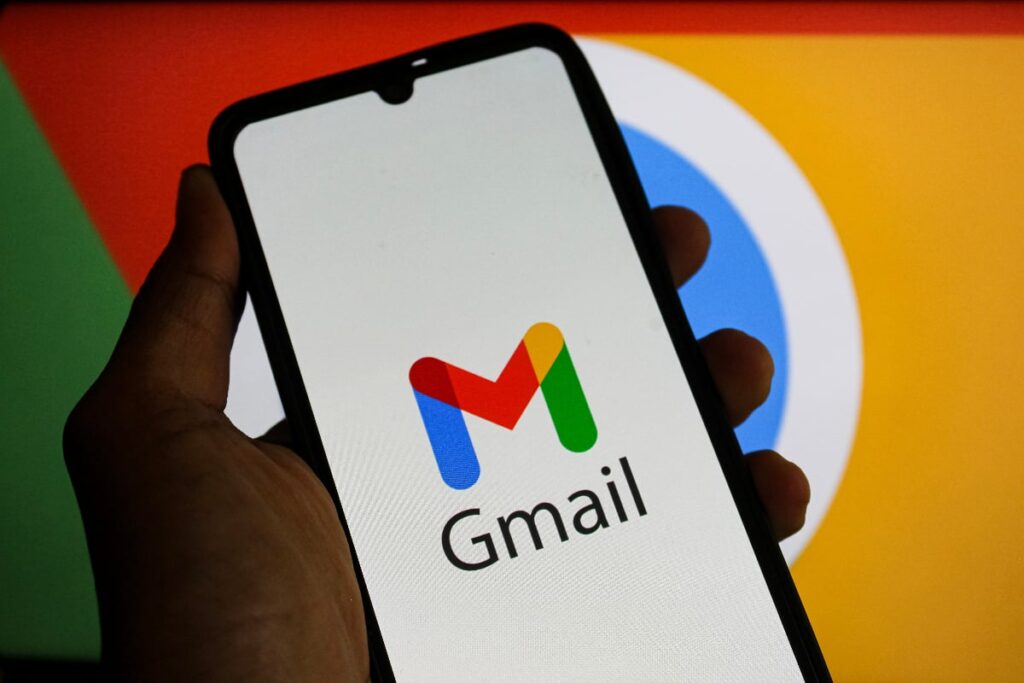 183 de milioane de parole furate. Cum să verifici dacă contul tău Gmail a fost compromis Imaginea prezintă un smartphone ținut în mână, pe al cărui ecran este afișată sigla serviciului de email Gmail (litera "M" stilizată în culorile roșu, albastru, verde și galben) împreună cu numele. Pe fundalul estompat se pot distinge culorile vii și elementele grafice specifice altor produse Google, subliniind asocierea aplicației cu ecosistemul companiei.
