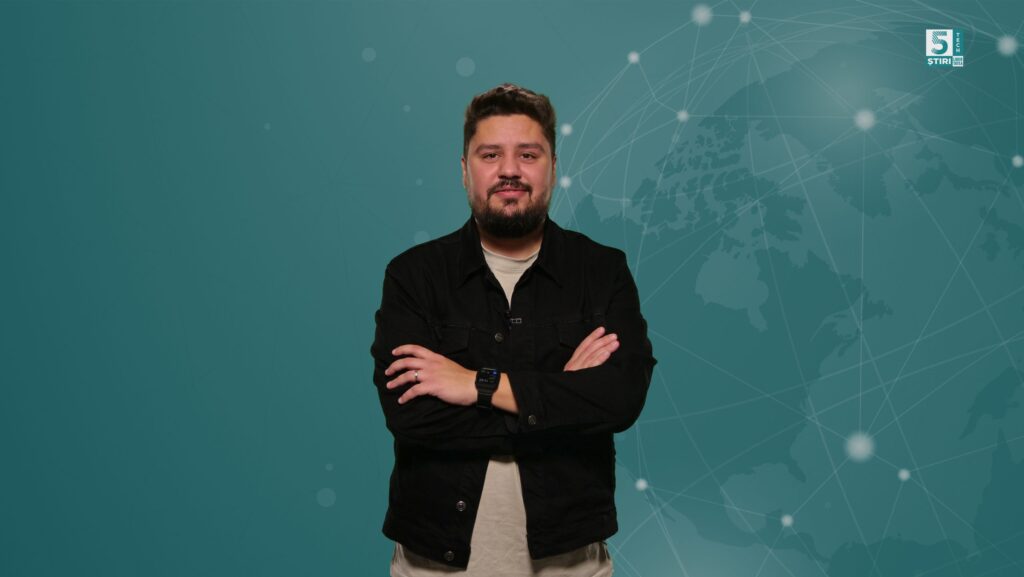 Claudiu Râpan, gazda rubricii 5 știri by Libertatea - Tech, cu un tricou bej și o cămașă neagră, cu mâinile încrucișate la piept, pe un fundal verde.