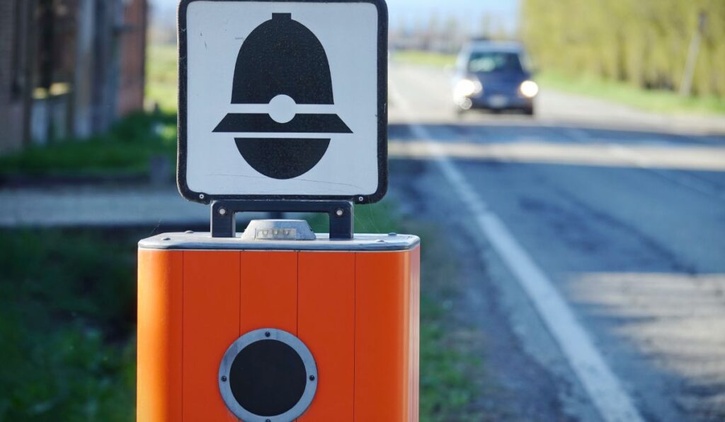 Cameră-radar din Italia, de culoare portocalie, pe marginea unui drum, cu un indicator rutier deasupra care arată o pălărie de carabinier
