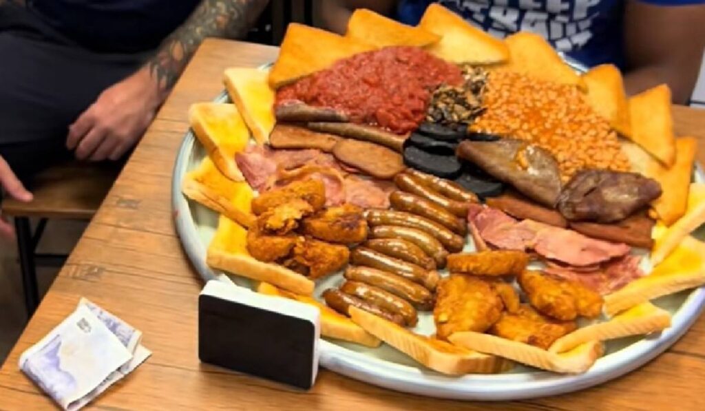 Un patou urias cu mancare de tip fast food, printre care 10 cârnați, mai multe felii de bacon, ciuperci, roșii, fasole, budincă neagră, opt cartofi prăjiți și 16 felii de pâine prăjită, asezat pe o mas[