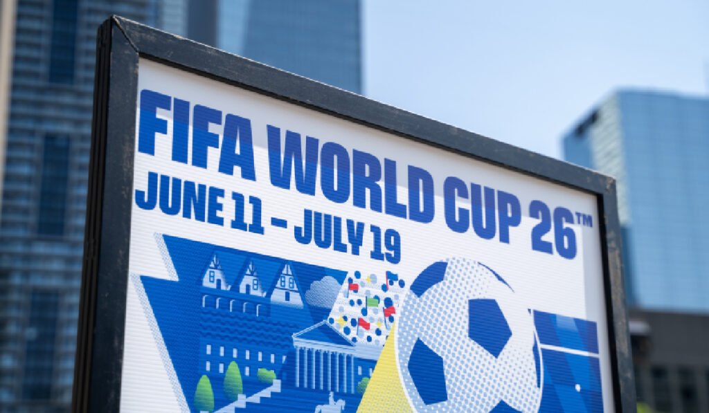 Imaginea prezintă de aproape un panou publicitar care anunță evenimentul sportiv FIFA World Cup '26 (Cupa Mondială 2026), specificând datele de desfășurare: 11 iunie - 19 iulie. Pe panou se află și o reprezentare grafică stilizată, incluzând o minge de fotbal și clădiri, iar în fundal se observă clădiri moderne de birouri.