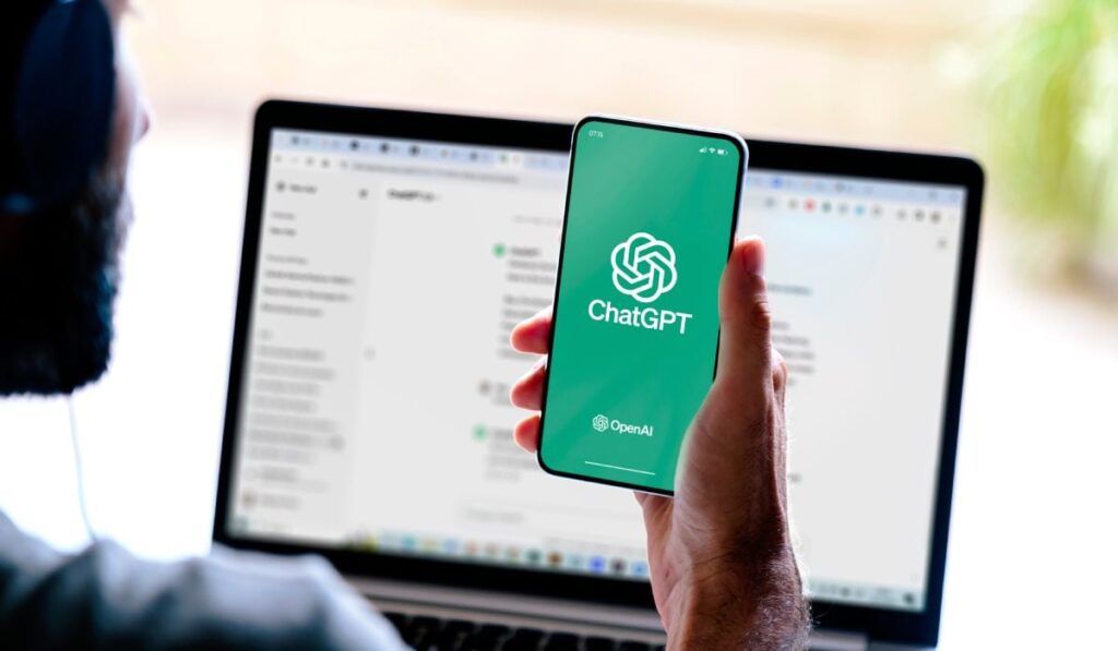 O persoană care ține în mână un telefon pe care apare logo-ul ChatGPT, pe fundal verde, iar pe fundal se vede în blur un laptop aprins