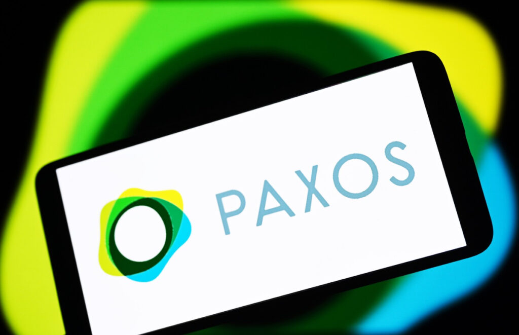 Imaginea arată sigla companiei de infrastructură blockchain Paxos pe ecranul unui smartphone, pe un fundal estompat și colorat. Sigla combină un simbol grafic modern, format din forme suprapuse de culoare galben, verde și albastru, cu numele "PAXOS" scris cu litere simple și subțiri de culoare albastru deschis.