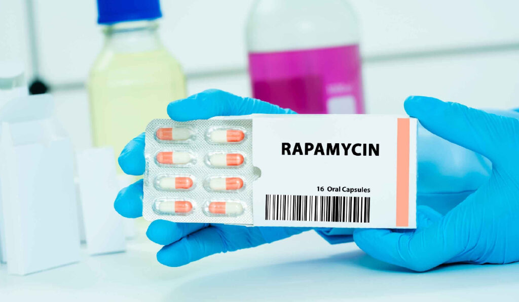 Imaginea prezintă de aproape o cutie de medicamente pe care este vizibil numele RAPAMYCIN și specificația "16 Oral Capsules", împreună cu un blister conținând capsule bicolore (alb și portocaliu). Mâini protejate de mănuși medicale albastre țin cutia, sugerând un mediu de laborator sau farmaceutic.