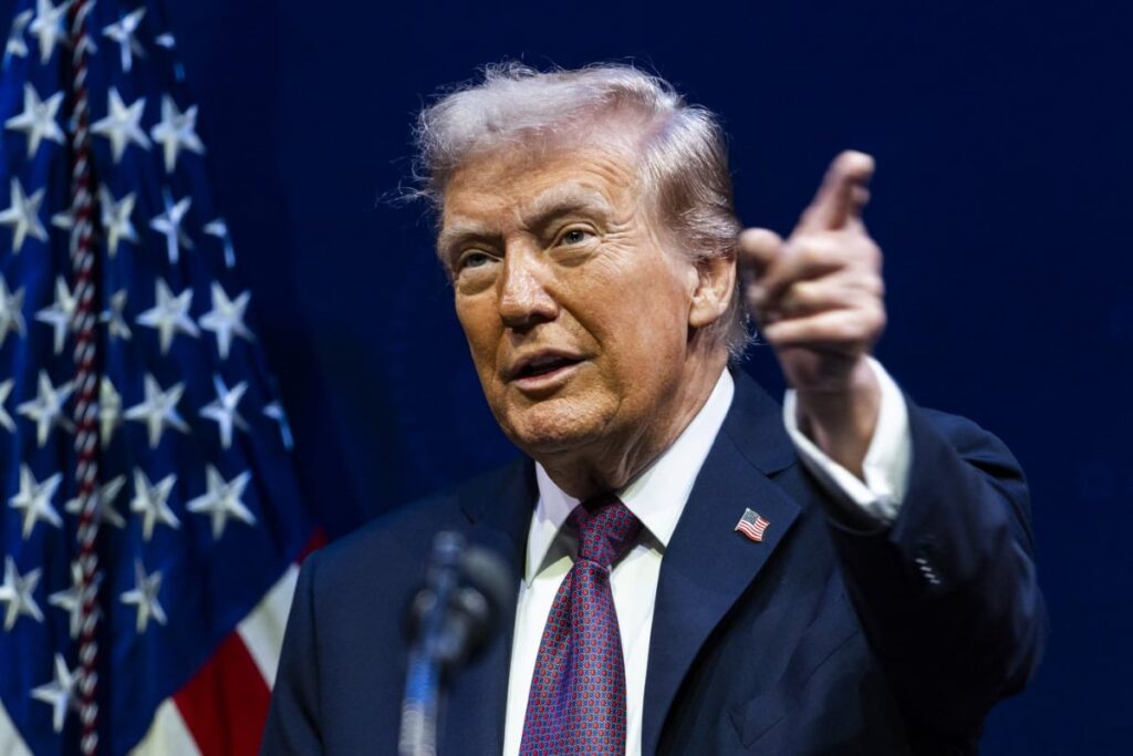 Imaginea îl prezintă pe Donald Trump vorbind la un eveniment, îmbrăcat într-un costum bleumarin, cămașă albă și o cravată roșie cu model. În timp ce vorbește, el ridică mâna dreaptă și arată cu degetul, având o expresie facială concentrată. În spatele său se poate observa un steag american.