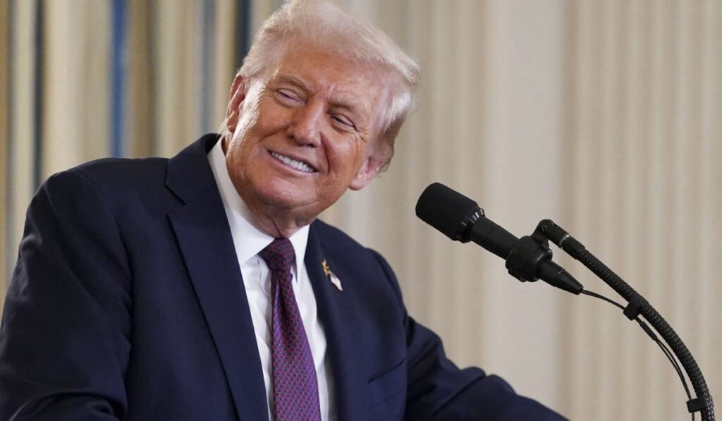 Președintele american Donald Trump, în timpul unei conferințe de presă la Casa Albă, poartă un costum negru și o cravată violet și zâmbește în fața camerei