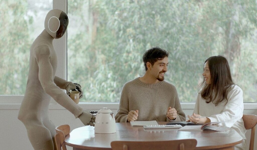 Un robot umanoid ajută un cuplu în gospodărie. Se vede cum pune ceainicul pe masă, în timp ce un bărbat și o femeie stau la masă și discută