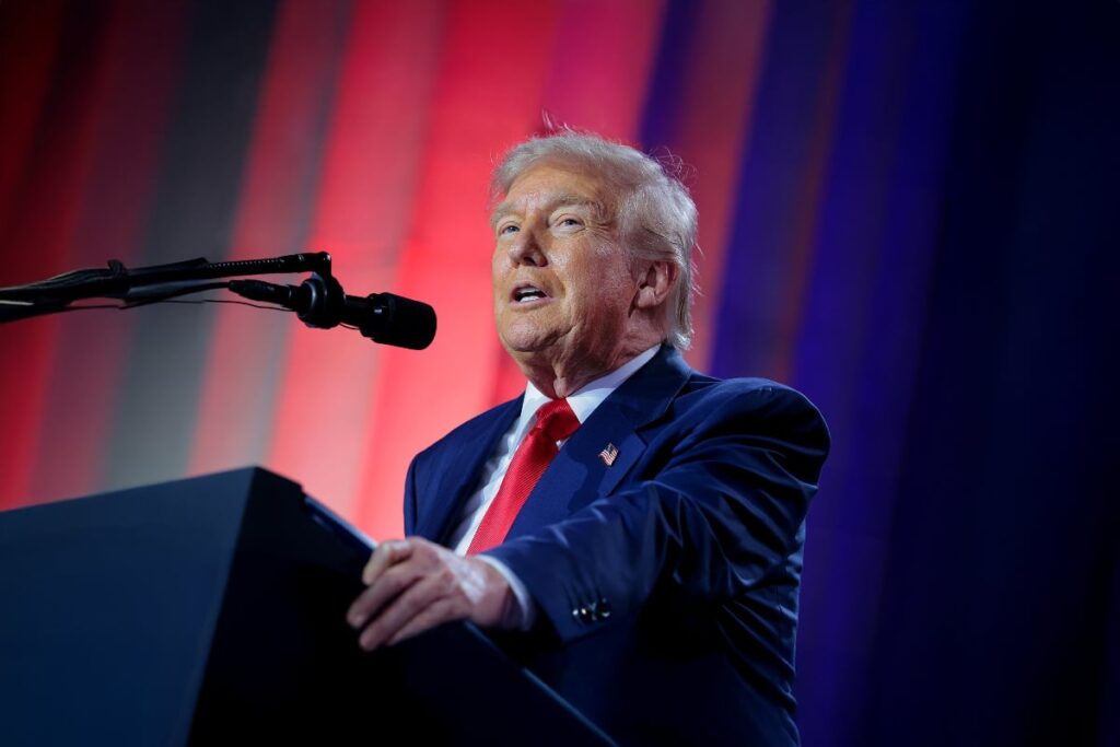 Cum îl oprește Constituția SUA pe Donald Trump din drumul spre un al treilea mandat Donald Trmup imbrăcat intr-un costum bleumarin, completat de o cravată roșie, vorbește la un pupitru în fața unui microfon, fiind luminat puternic. În fundal se observă o scenă cu iluminare dramatică în culorile roșu, alb și albastru, sugerând că este un eveniment politic, un discurs sau o conferință.