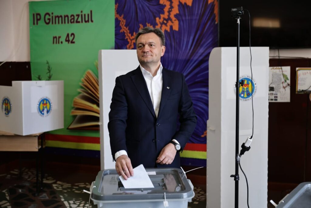 Imaginea îl surprinde pe Dorin Recean, Prim-ministrul Republicii Moldova, îmbrăcat în costum, în timp ce introduce un buletin de vot în urna transparentă. Scena are loc într-o secție de votare, având în fundal un afiș verde care menționează "IP Gimnaziul nr. 42" și un panou cu simbolul de stat al Republicii Moldova.