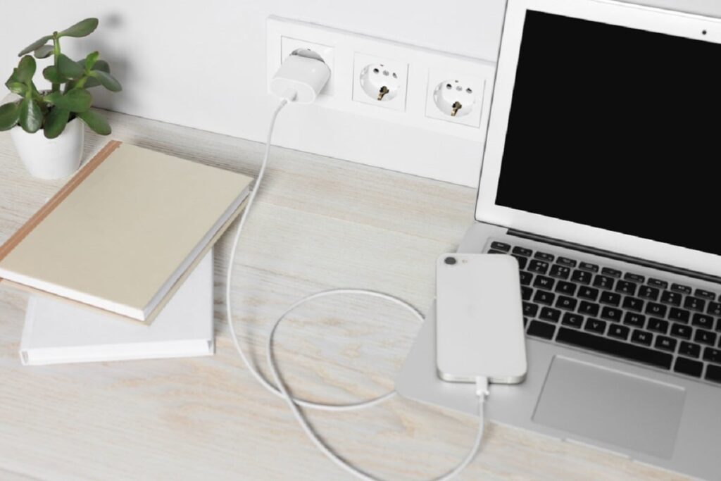 De ce se încarcă mai greu telefonul tău? Cauze și soluții simple - Imagine cu un iPhone pus la încărcat şi aşezat pe tastatura unui laptop, alături de două agende şi de o mică plantă verde într-un ghiveci alb, toate pe o masă albă.