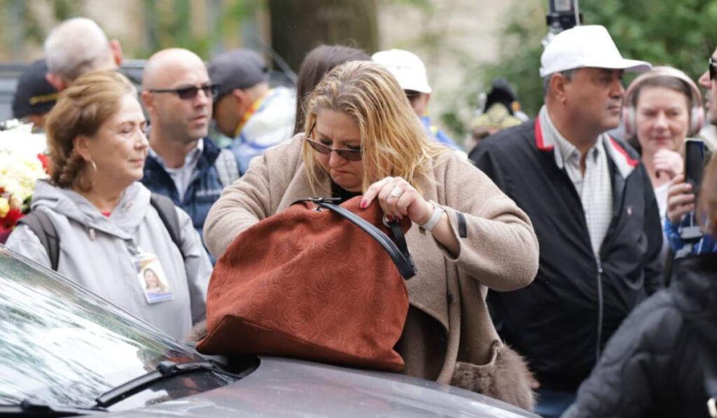 Diana Șoșoacă, în timp ce își caută telefonul mobil în geantă. Poșeta maro este așezată pe capota unei mașini ge jandarmi, iar europarlamentarul are ochelari de soare la ochi și o expresie dezamăgita