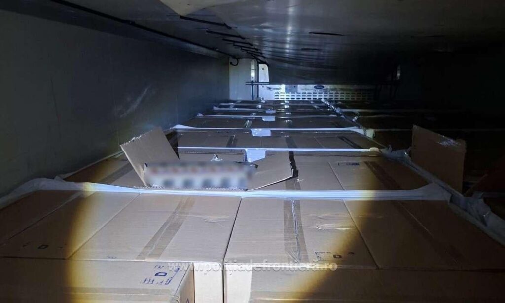 Imaginea arată interiorul unui spațiu restrâns, probabil o remorcă de camion sau un container, umplut de jos în sus cu numeroase cutii de carton mari, ambalate. Un spațiu îngust este lăsat liber, permițând vizualizarea plafonului metalic și a rândurilor de cutii care se extind în adâncime.