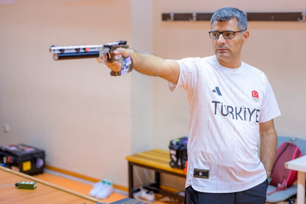 Imaginea îl surprinde pe Yusuf Dikeç, un sportiv turc, concentrat în timpul unui antrenament de tir, țintind cu un pistol sportiv cu aer comprimat. El poartă un tricou alb cu inscripția TÜRKİYE (Turcia) și simbolurile olimpice, într-un poligon de tragere interior.