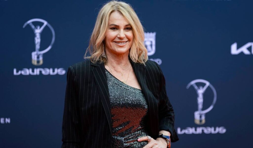Gimnasta Nadia Comăneci, pe covorul roșu, îmbrăcată într-o salopetă argintie cu un sacou negru din catifea. Ține o mână în talie și privește fix în fața camerei