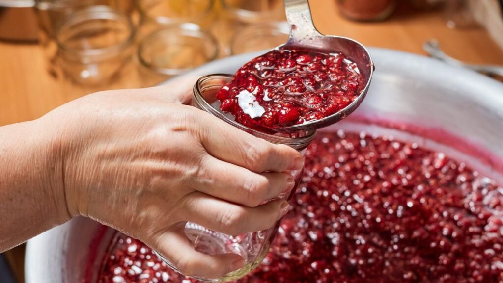 O femeie toarnă dulceață de zmeură proaspăt preparată acasă într-un borcan de sticlă folosind o lingură de bucătărie.