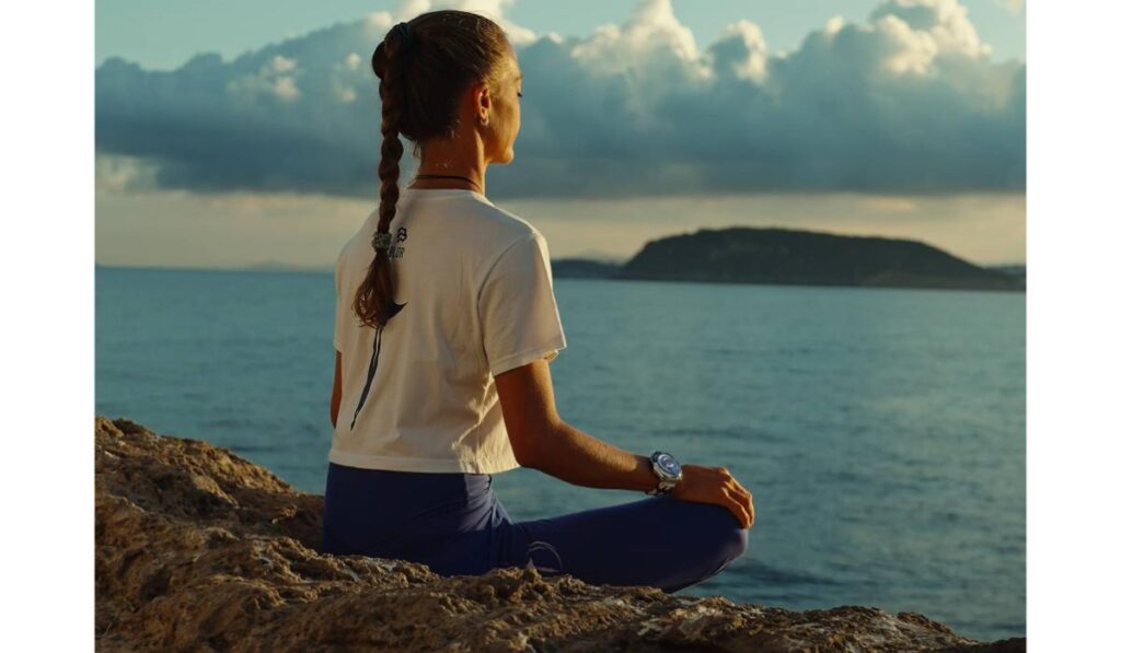 Huawei - poza cu o femeie meditanad la malul oceanului. Are parul saten, prins intr-o coada impletita, sta cu picioarele incrucisate si poarta un tricoul alb si colanti bleumarin