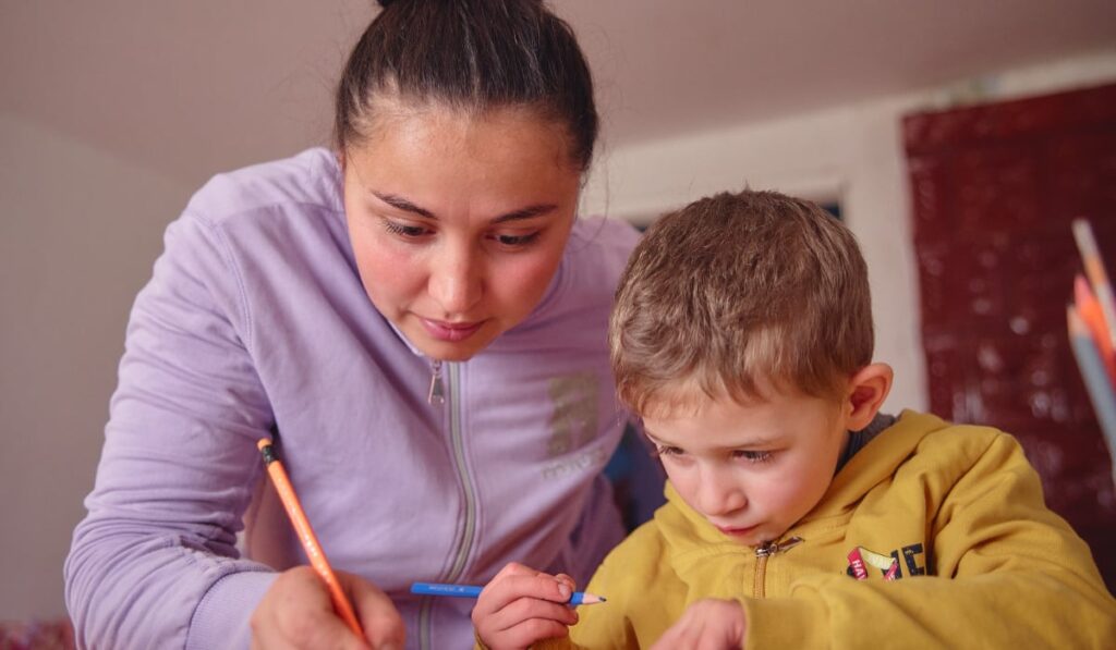 O mamă și fiul ei care sunt aplecați peste o masă și lucrează împreună la lecții. Amândoi folosesc creioane colorate, mama scriind pe hârtie în timp ce îl ghidează pe copil.
