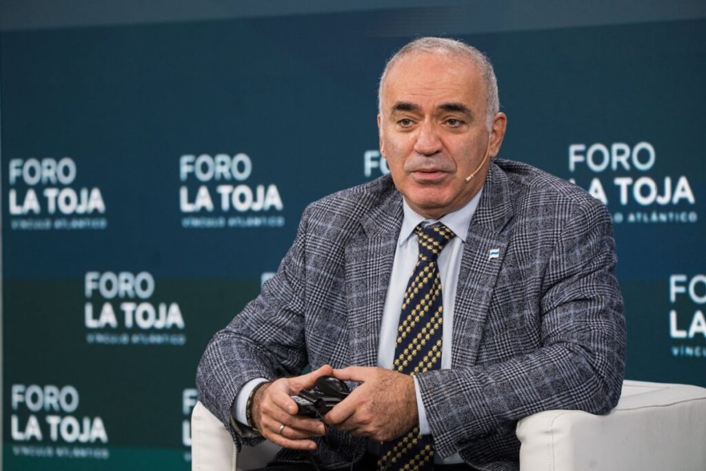 Imaginea îl înfățișează pe Garry Kasparov, fost campion mondial la șah, stând pe un scaun alb și purtând un sacou în carouri gri și o cravată cu model în dungi galbene și albastre. El participă la un eveniment, având în spate un panou cu sigla „FORO LA TOJA” (Forumul La Toja), care indică contextul apariției sale publice.