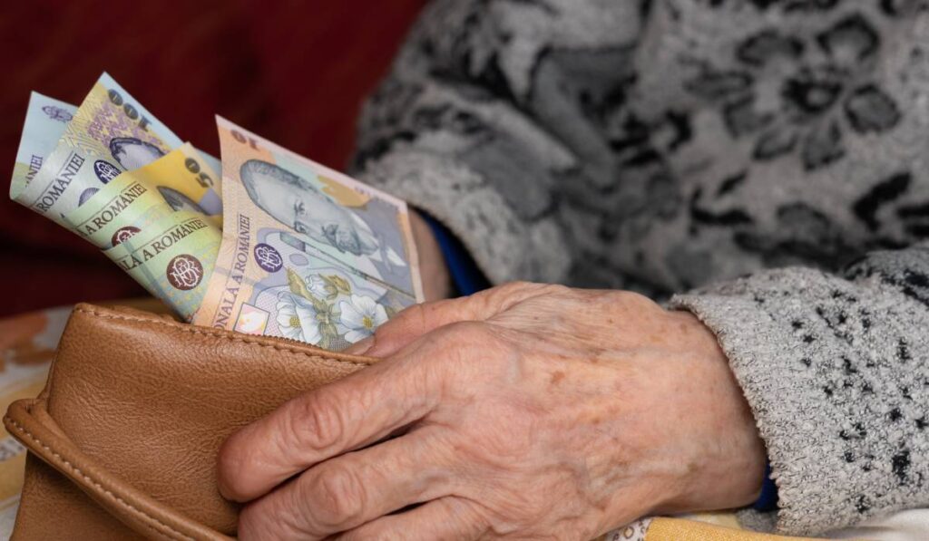 O femeie în vârstă care stă jos și își numără banii din portofel. Femeia poartă un pulover gri cu un model, portofelul pe care îl ține în mână este din piele maro, iar din portofel ies bancnote de 100, 50 și 10 lei.