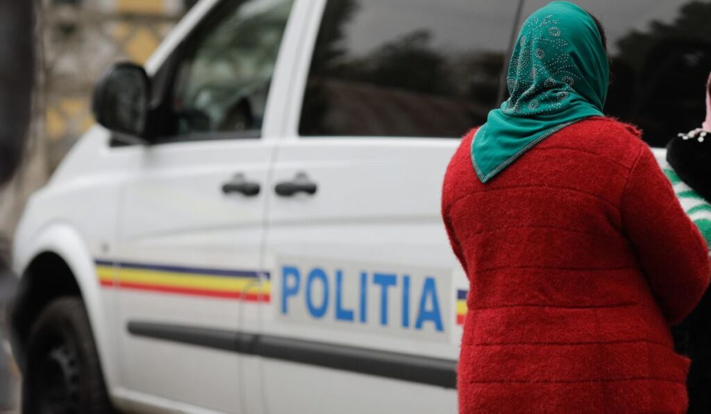 Femeie îmbrăcată cu un pulover roșu, cu batic verde pe cap, lângă o mașină de Poliție