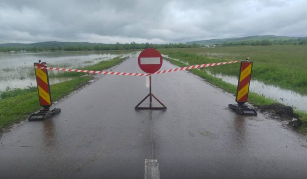 Imaginea surprinde un drum județean complet închis circulației, marcat cu bandă de interdicție și semn de acces interzis, din cauza inundațiilor.