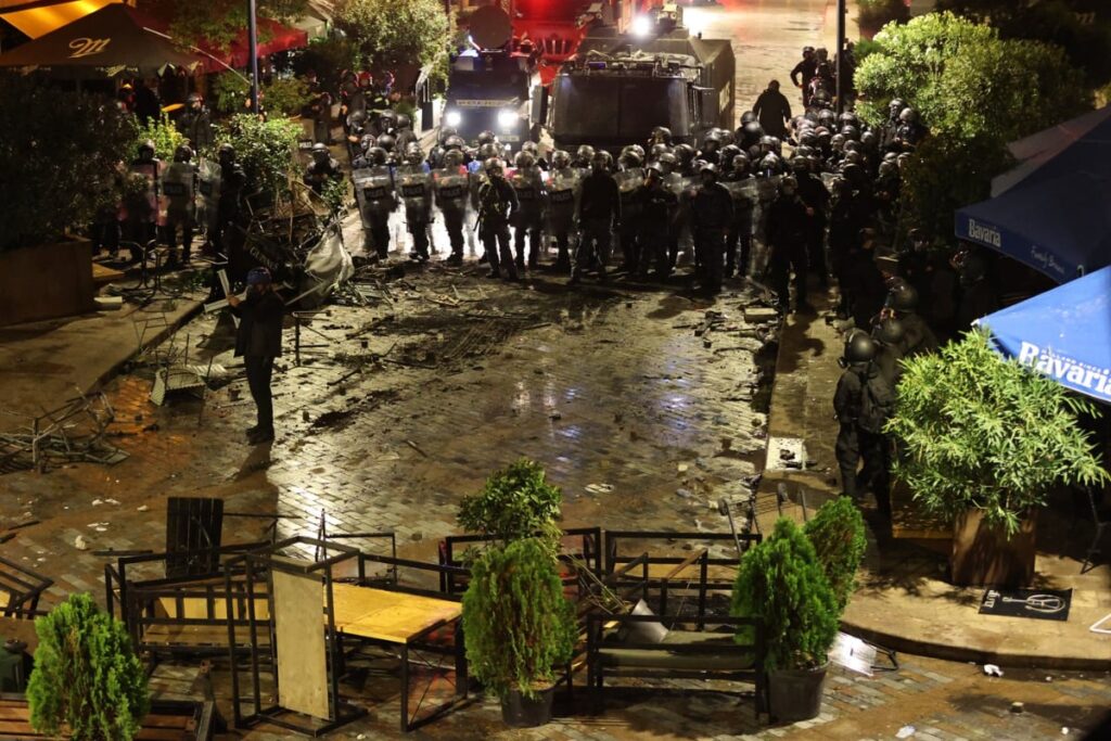 Imaginea surprinde o scenă de tensiune nocturnă, unde un rând compact de forțe de ordine (posibil poliție sau jandarmerie) în echipament de intervenție se confruntă cu o zonă dezordonată și plină de resturi. În prim-plan se văd mese și scaune răsturnate, iar pe fundal, două vehicule blindate și un grup mare de agenți menținând o linie de apărare, sugerând o intervenție după tulburări civile sau un protest.