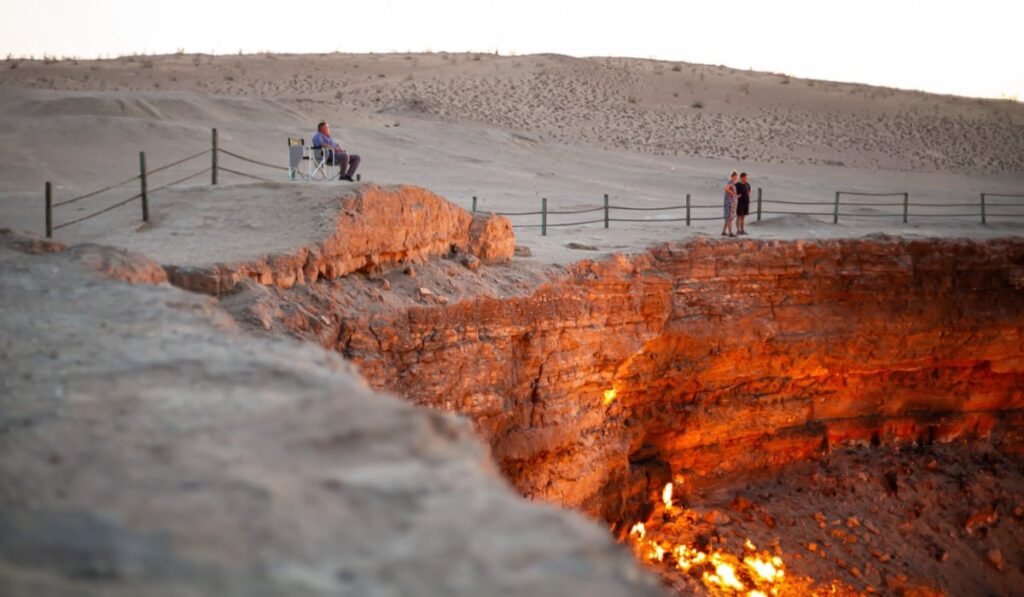 Șapte dintre cele mai neobișnuite destinații turistice din lume Imaginea prezintă craterul Darvaza, iar pe margine sunt turiști care privesc flăcările.