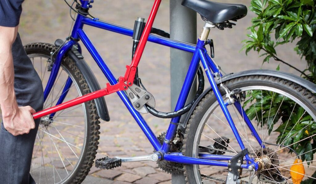 În imagine se vede o bicicletă de culoare albastră, sprijinită de un stâlp gri. Bicicleta are un lanț negru pe cadru, iar în prim plan apare o mână ținând un clește roșu, care este poziționat pe lanț.