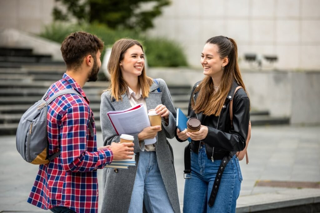Unde aleg tot mai mulți tineri români să studieze. Țara despre care spun că le oferă „multe oportunități” Imaginea surprinde trei studenți tineri care stau de vorbă și zâmbesc, ținând în mâini caiete și cești de cafea de carton, sugerând o pauză între cursuri. Ei sunt îmbrăcați casual, în haine moderne (blugi, sacouri și o jachetă de piele), cu fundalul exterior sugerând un campus universitar.
