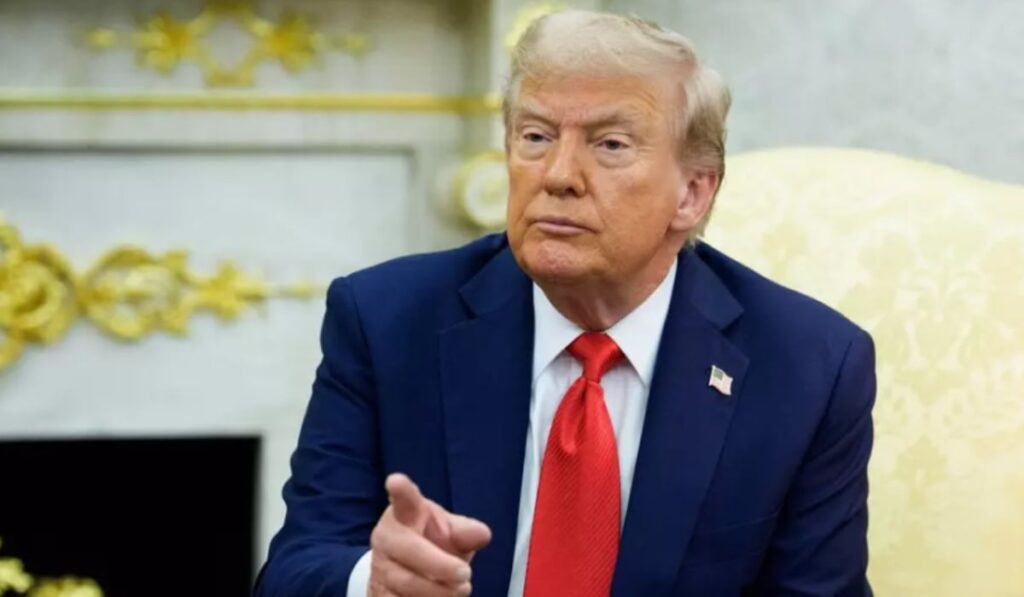 Donald Trump în sacou negru, cămașă albă și cravată roșie, în timp ce stă pe scaun