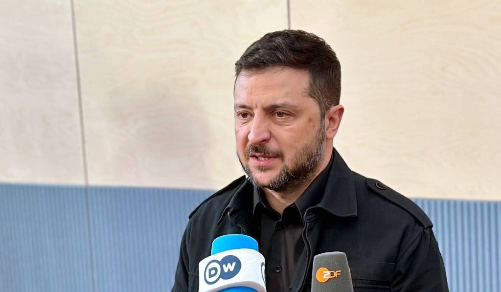 Volodimir Zelenski vorbește cu presa, în fața sa sunt câteva microfoane și este îmbrăcat în negru