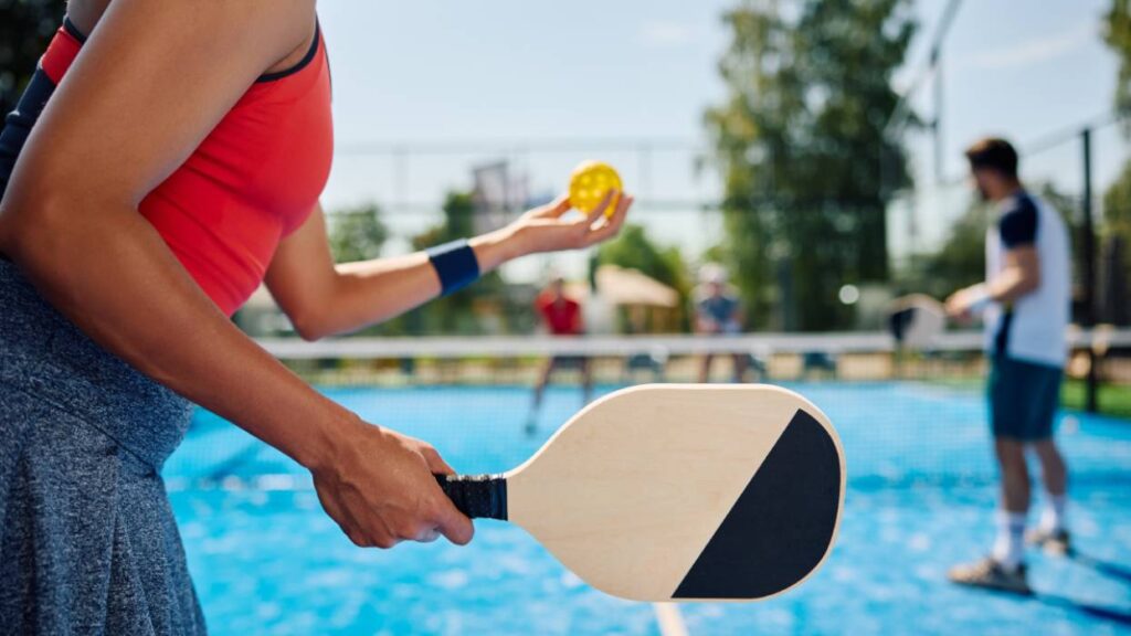 O femeie pe un teren de tenis, in mana stanga cu o minge mica, galbena, cu gauri si in mana dreapta o paleta de ping-pong, in findal se vad estompati alti cocurenti intr-un meci de pickleball.